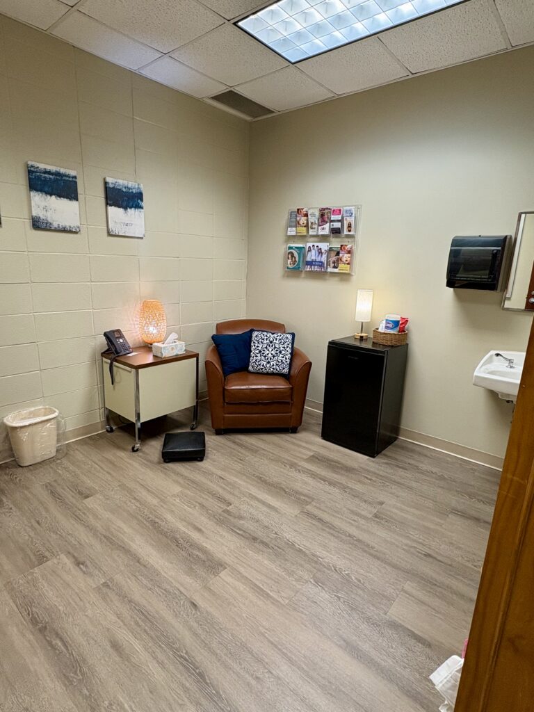 Lactation room with two brown leather chairs, a small side table with a lamp and tissues, a mini refrigerator, wall-mounted literature rack, sink, and wood-look flooring.