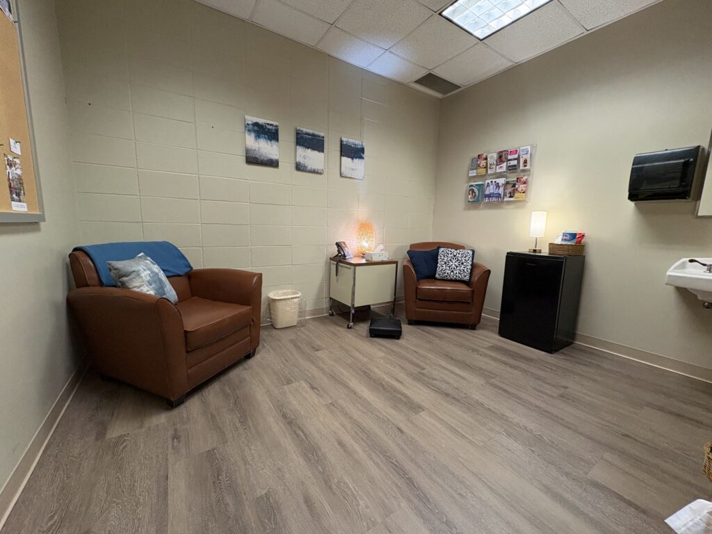 Lactation room with two brown leather chairs, a small side table with a lamp and tissues, a mini refrigerator, wall-mounted literature rack, sink, and wood-look flooring.