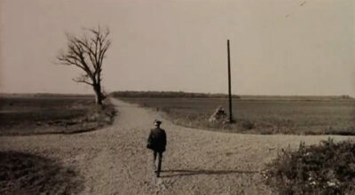 Man walking on dirt road, approaching a four way intersection. There is a telephone or telegraph line pole on the right and a leafless tree on the left. Sepia tone. 