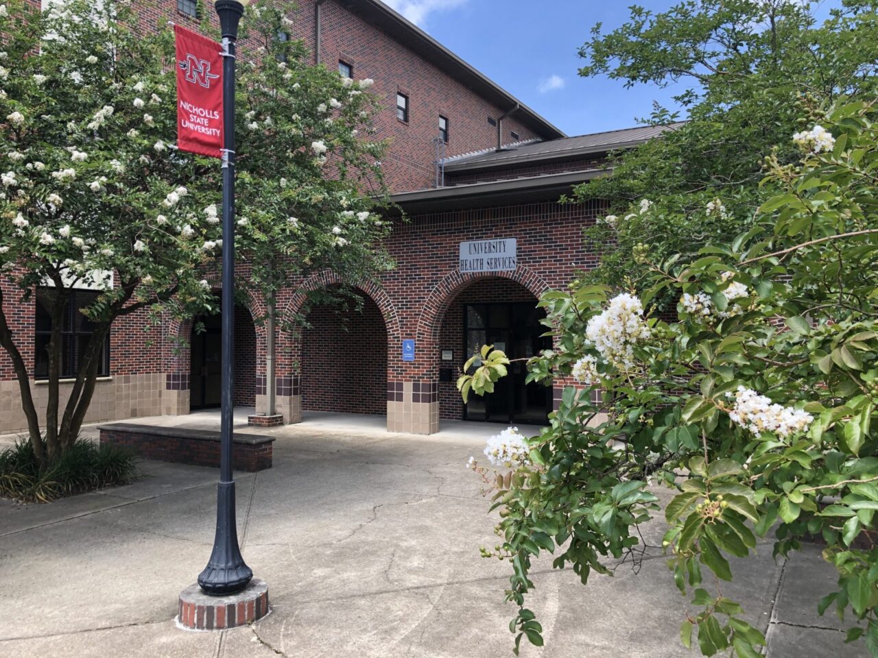 Exterior of Nicholls State University’s University Health Services building, featuring a brick facade, arched entrances, campus banners, and flowering trees surrounding the walkway.