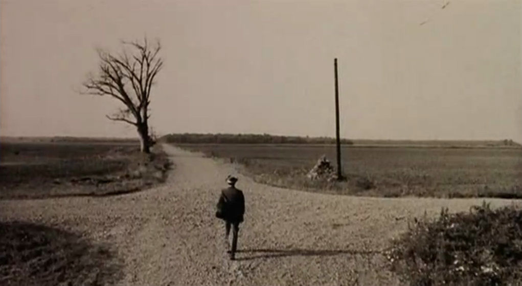 Man walking on dirt road, approaching a four way intersection. There is a telephone or telegraph line pole on the right and a leafless tree on the left. Sepia tone. 
