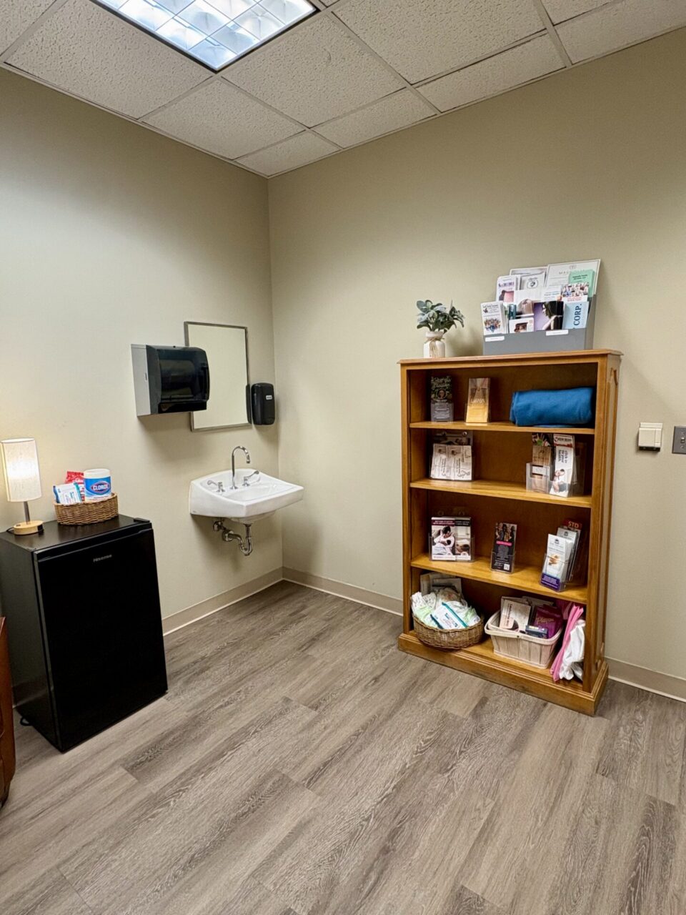 Lactation room with a mini refrigerator, bookshelf, sink, and wood-look flooring.